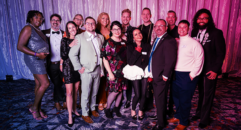 Group of professionally dressed individuals posing together at a formal event with a curtain backdrop and patterned carpet.