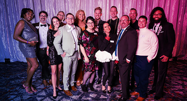 Group of professionally dressed individuals posing together at a formal event with a curtain backdrop and patterned carpet.